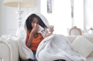 Lady wrapped in a blanket on the couch, sick, blowing her nose.